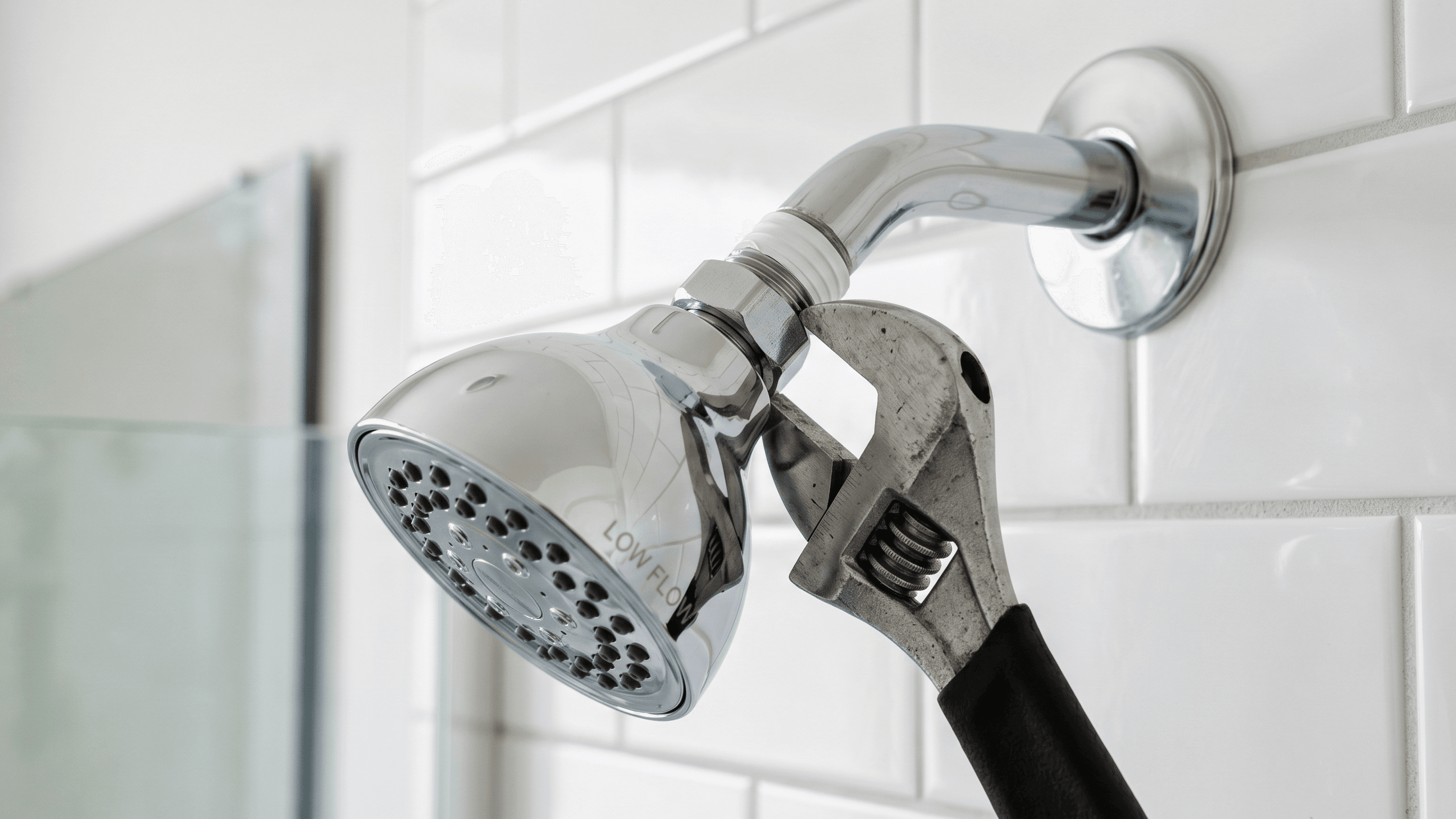 An adjustable wrench tightening a new chrome low-flow showerhead onto a shower arm with white plumber's tape, set against a clean white subway tile bathroom wall.